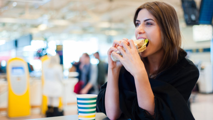 A woman in a black shirt eats a sandwich in an airport restaurant; Best airports for local food