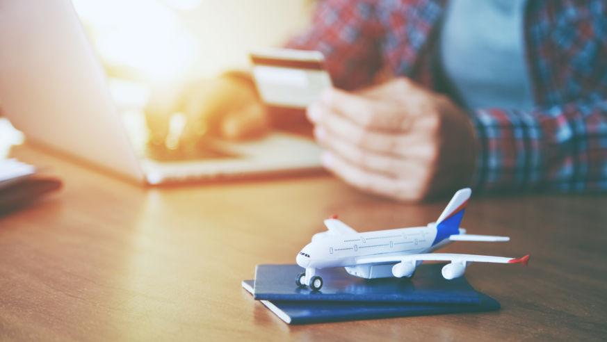 A man books a trip on a computer with his credit card. A toy airplane sits on a passport in the foreground