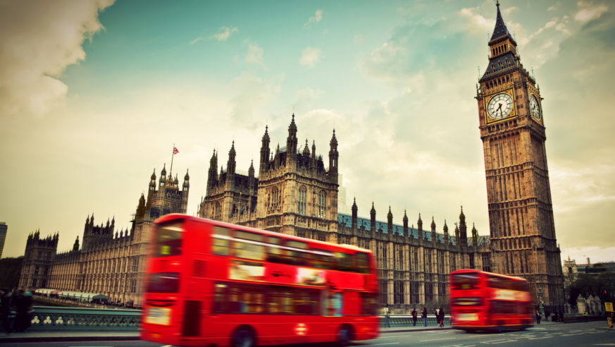 A street-level view of a London red bus with the Houses of Parliament behind it.