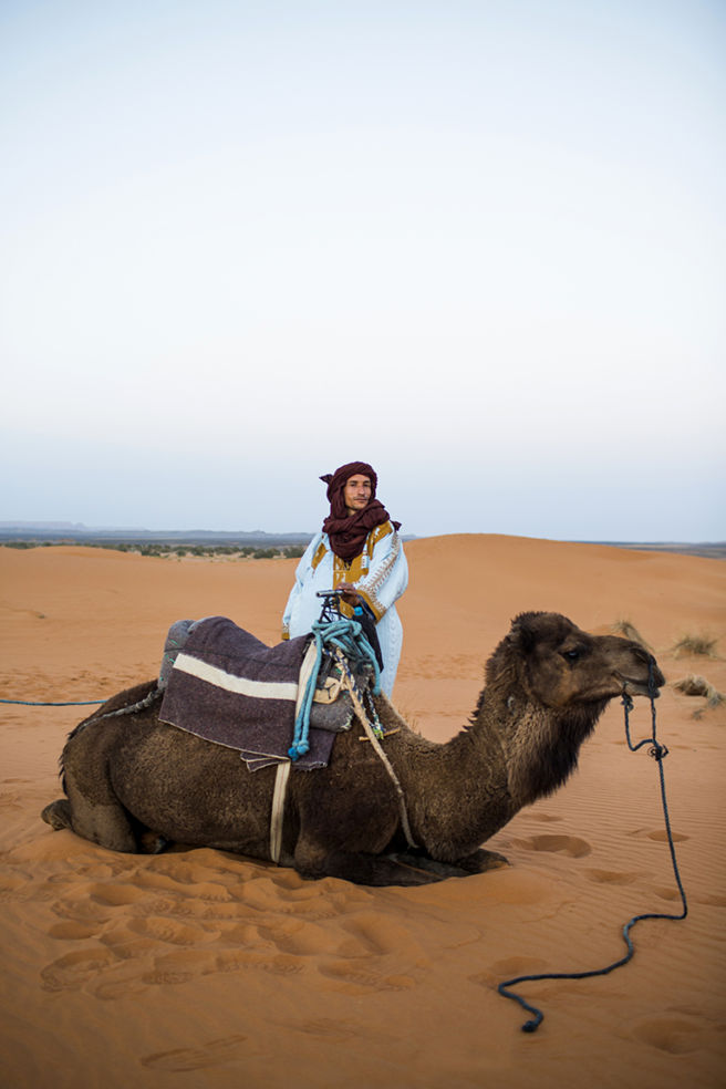 A guide stands by his camel as the soft light of sunset falls and the desert begins to cool